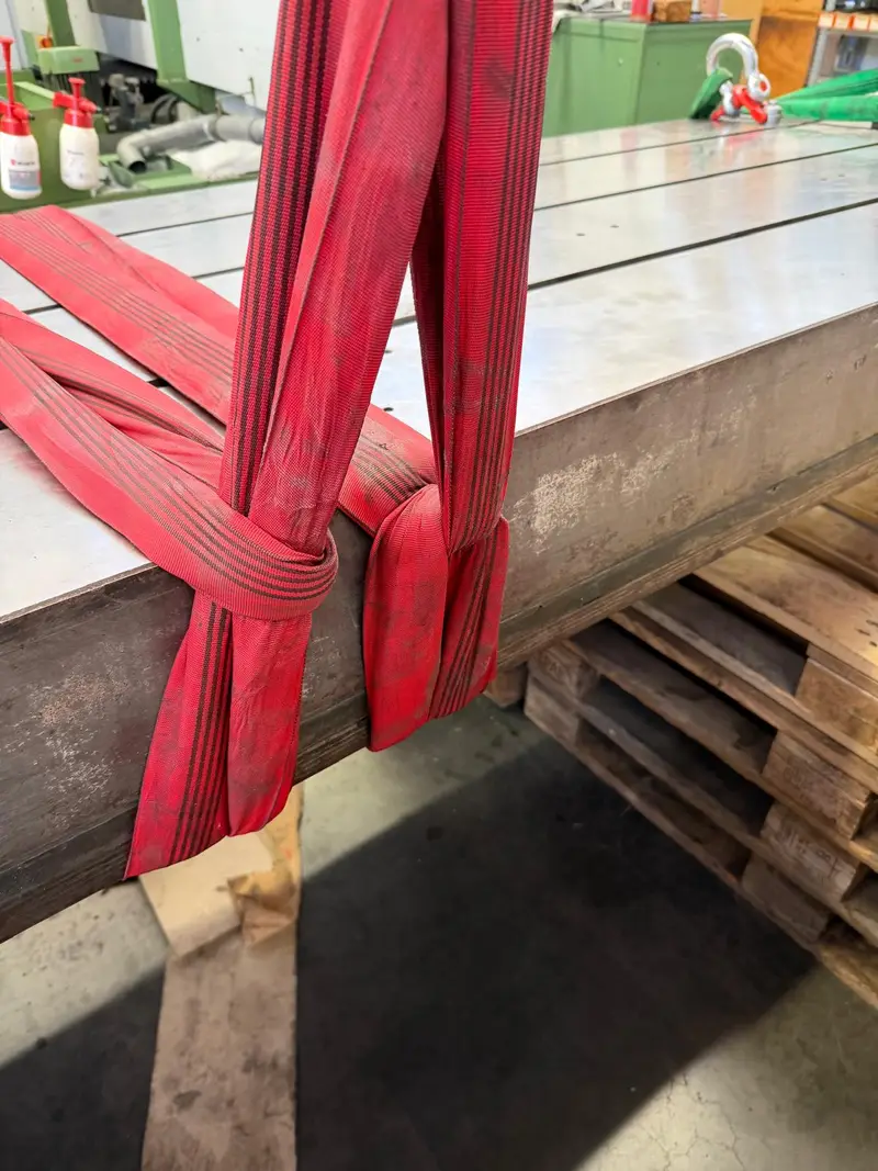 Eslinga roja alrededor de una pieza de máquina en metal desnudo, apoyada sobre palés.