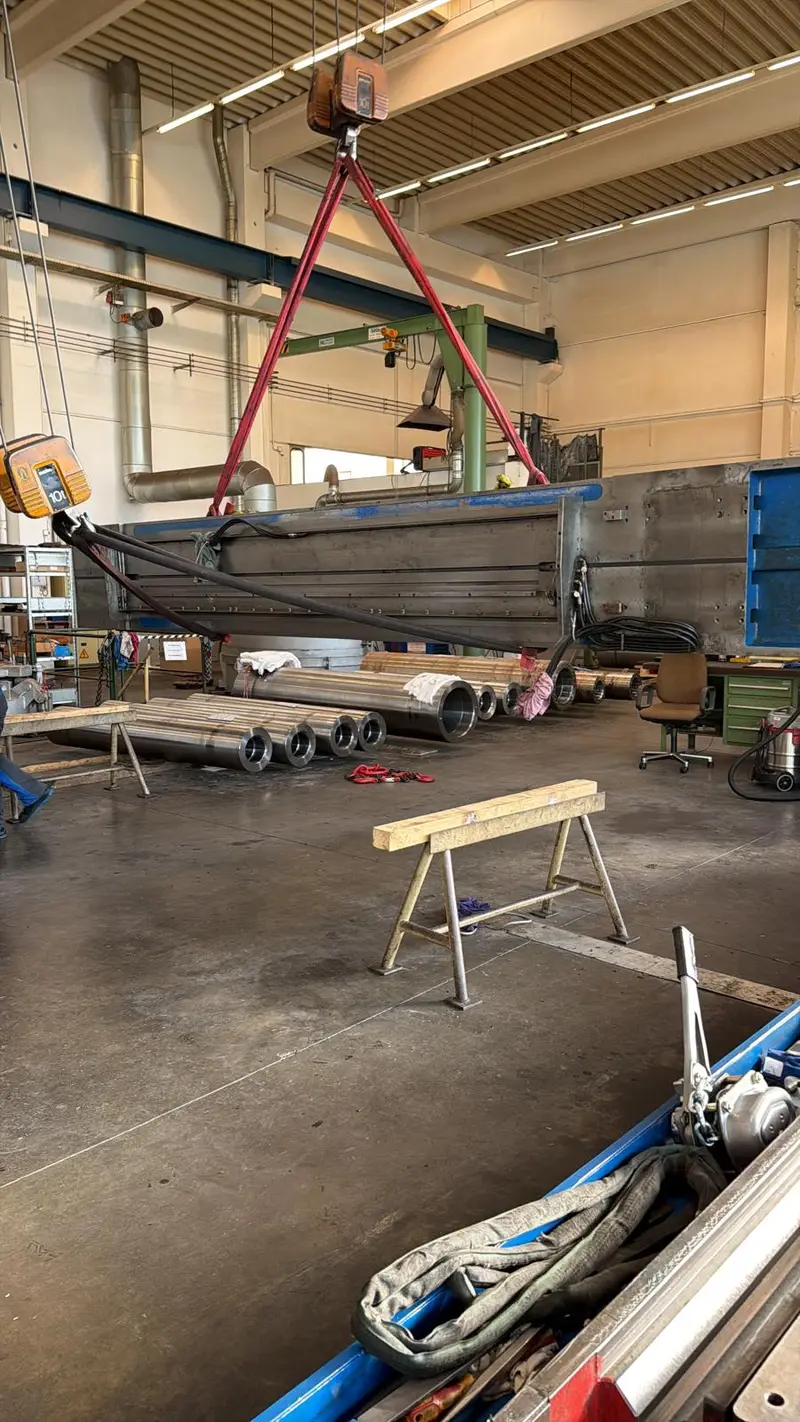 Bancada larga colgada de la grúa sobre el suelo de la nave; delante, un caballete de madera.