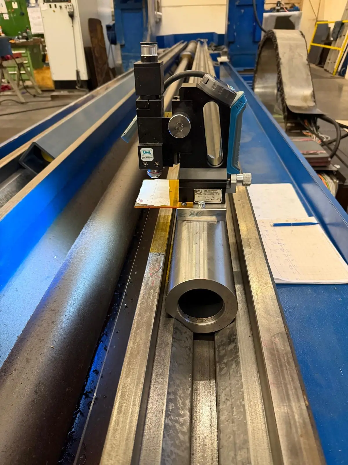 UHL FLZ alignment tester on a steel tube in the way of a long, blue-painted machine bed. Next to it lie a measuring sheet with handwritten readings and a pen.