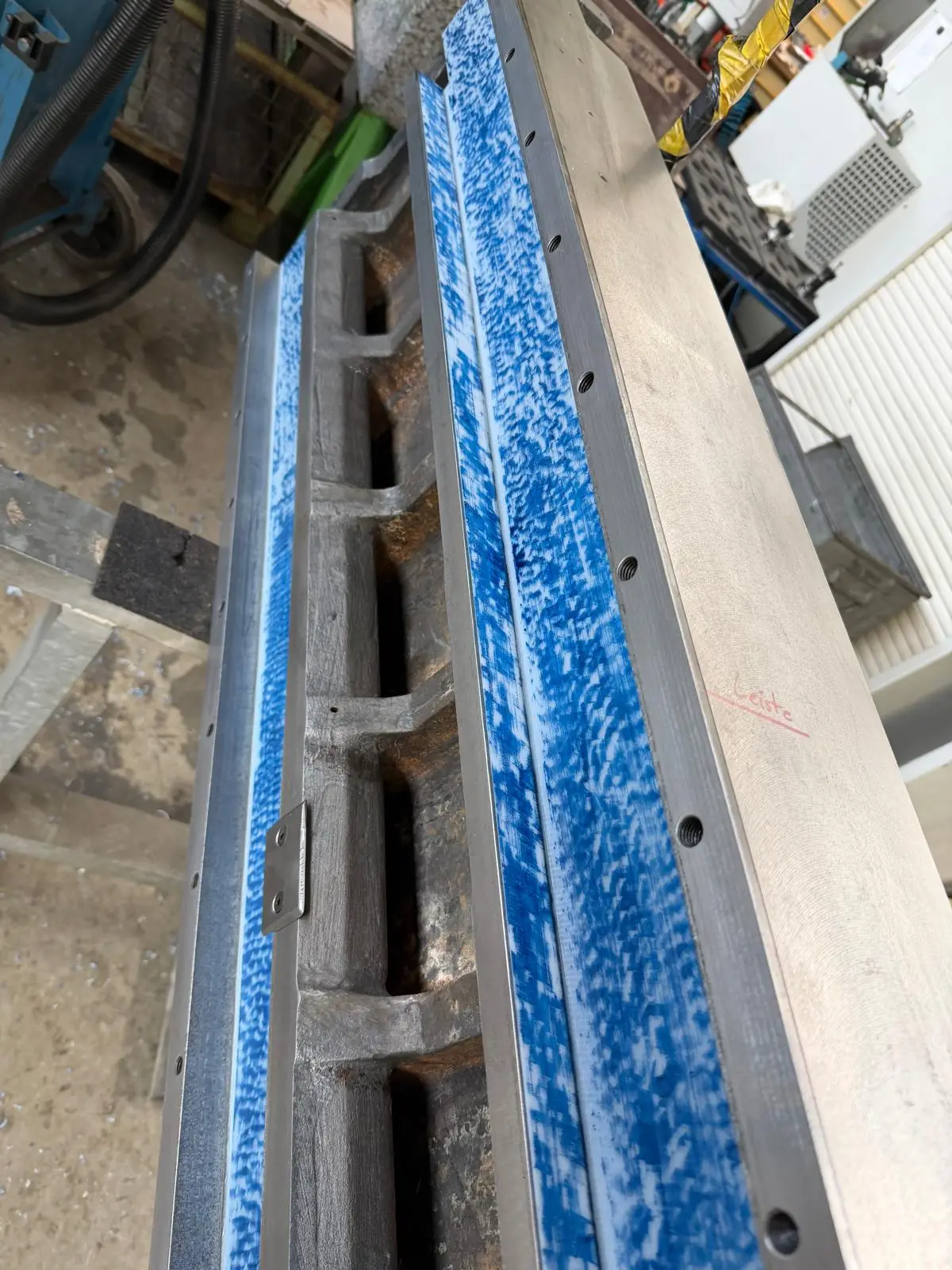 Two guide strips on a machine bed, spotted in blue, seen at an angle from above. The German word Leiste (strip) is written on the casting in red marker.
