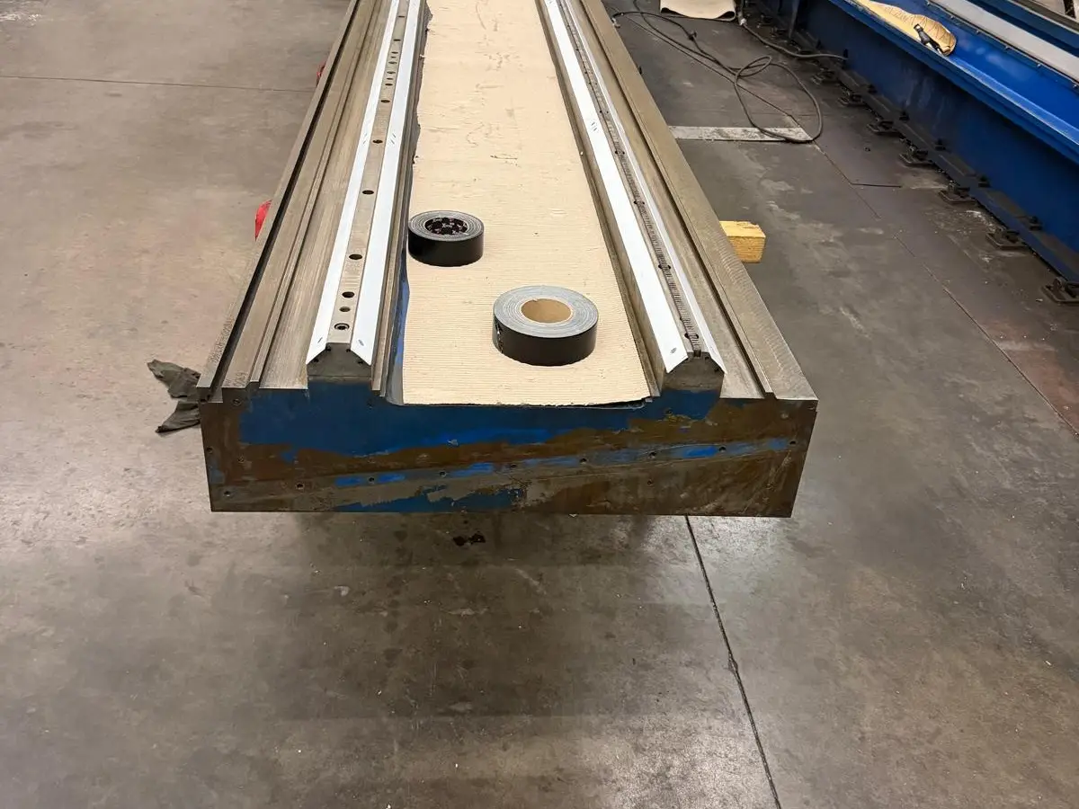 Long machine bed, out of the machine, on the shop floor, painted blue with rust on the end face. The center between the ways is lined with cardboard, with two rolls of tape on it.