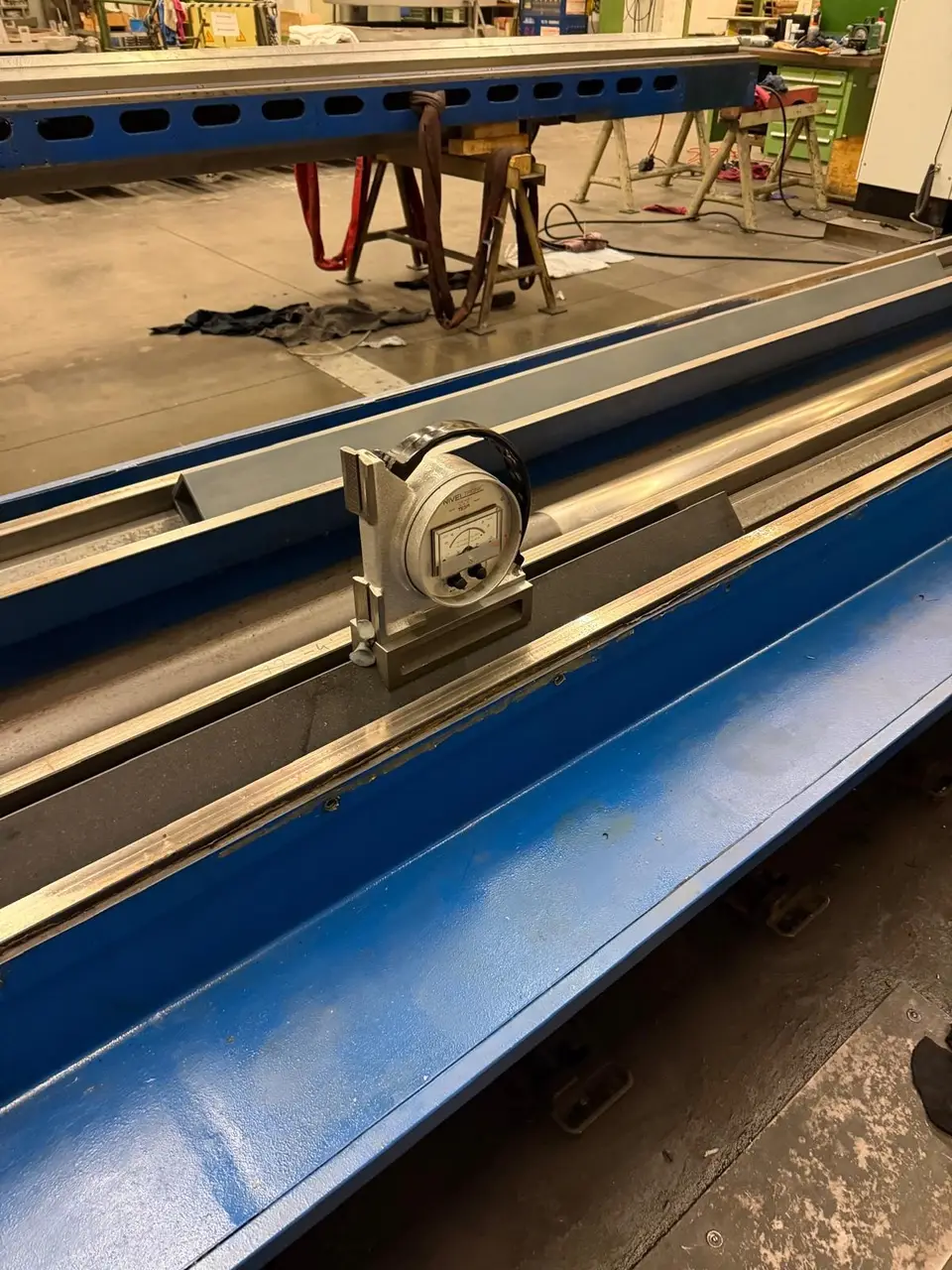 Nivel electrónico NIVELTRONIC de TESA en su soporte, sobre una regla oscura colocada en la guía de una bancada larga pintada de azul. Detrás, la nave con caballetes y eslingas.