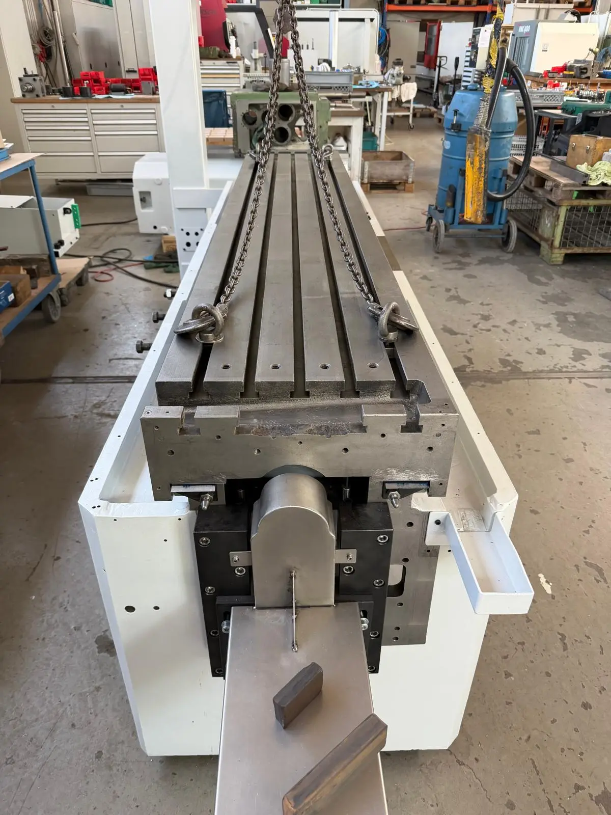 Machine bed painted white in the workshop. The table with its T-slots hangs from a chain sling and is being set onto the ways.
