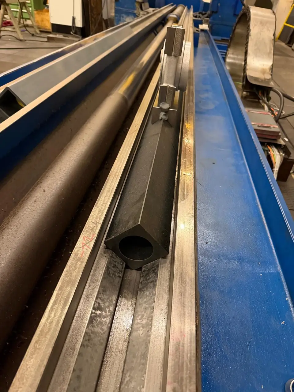 Regla cuadrada negra y hueca en la guía de una bancada larga pintada de azul. En el extremo, un soporte de aluminio con tornillos de ajuste.
